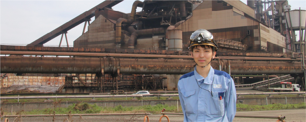 画像：大きな工場を背景に、作業着を着て、ゴーグルがついているヘルメットを被った若い男性作業員