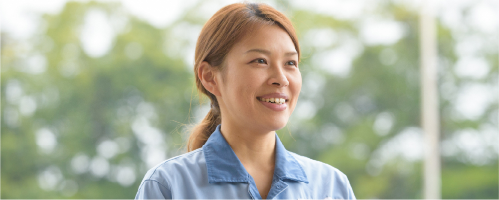 画像：緑がある場所を背景に、作業着をきた女性作業員。長い髪を後ろに束ねて、笑顔でいる様子